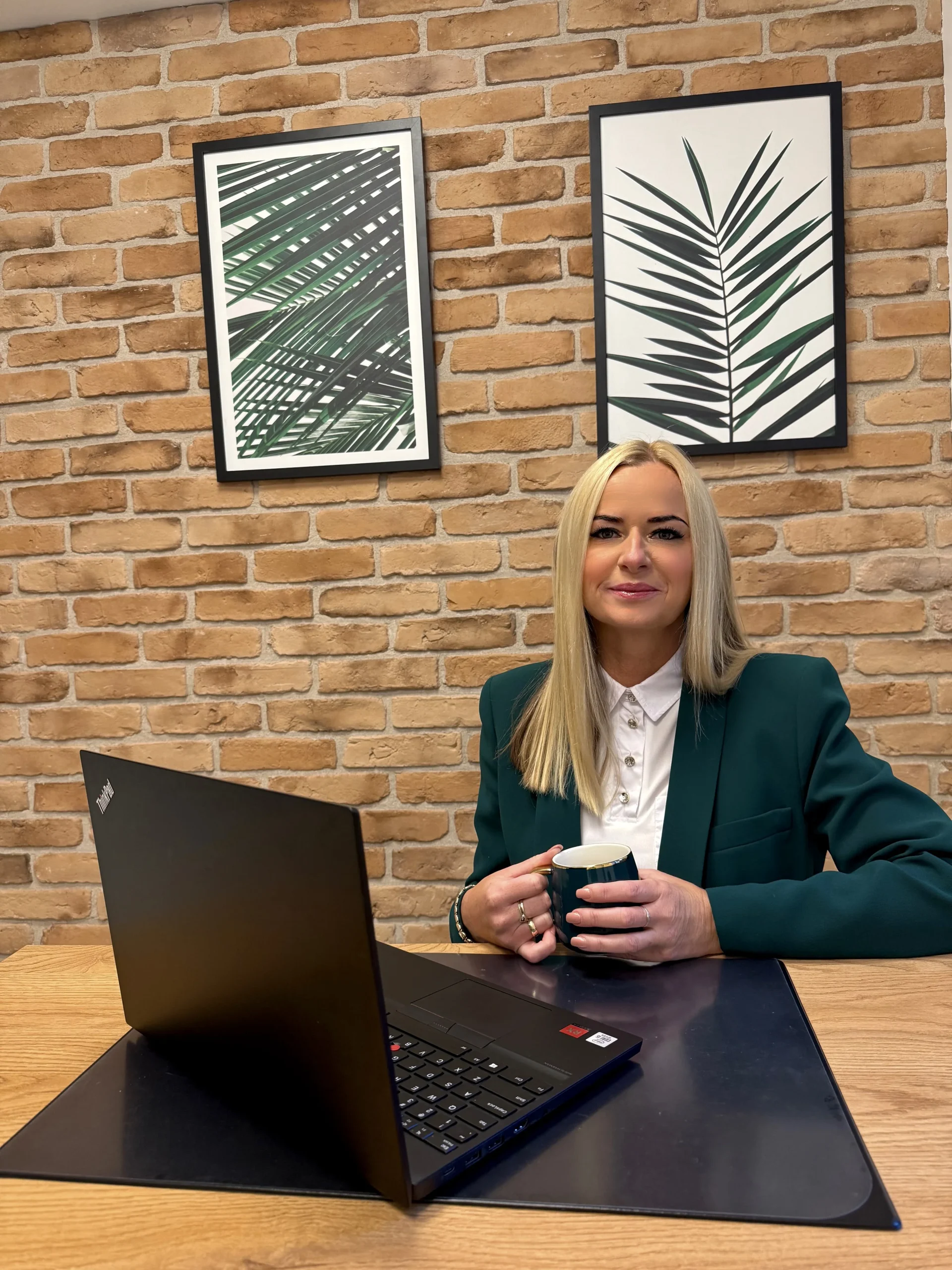 Uśmiechnięta kobieta z długimi, blond włosami siedzi przy biurku z laptopem, trzymając kubek. Ma na sobie zieloną marynarkę. W tle widać ścianę z czerwonej cegły i dwa oprawione obrazy z motywem liści.