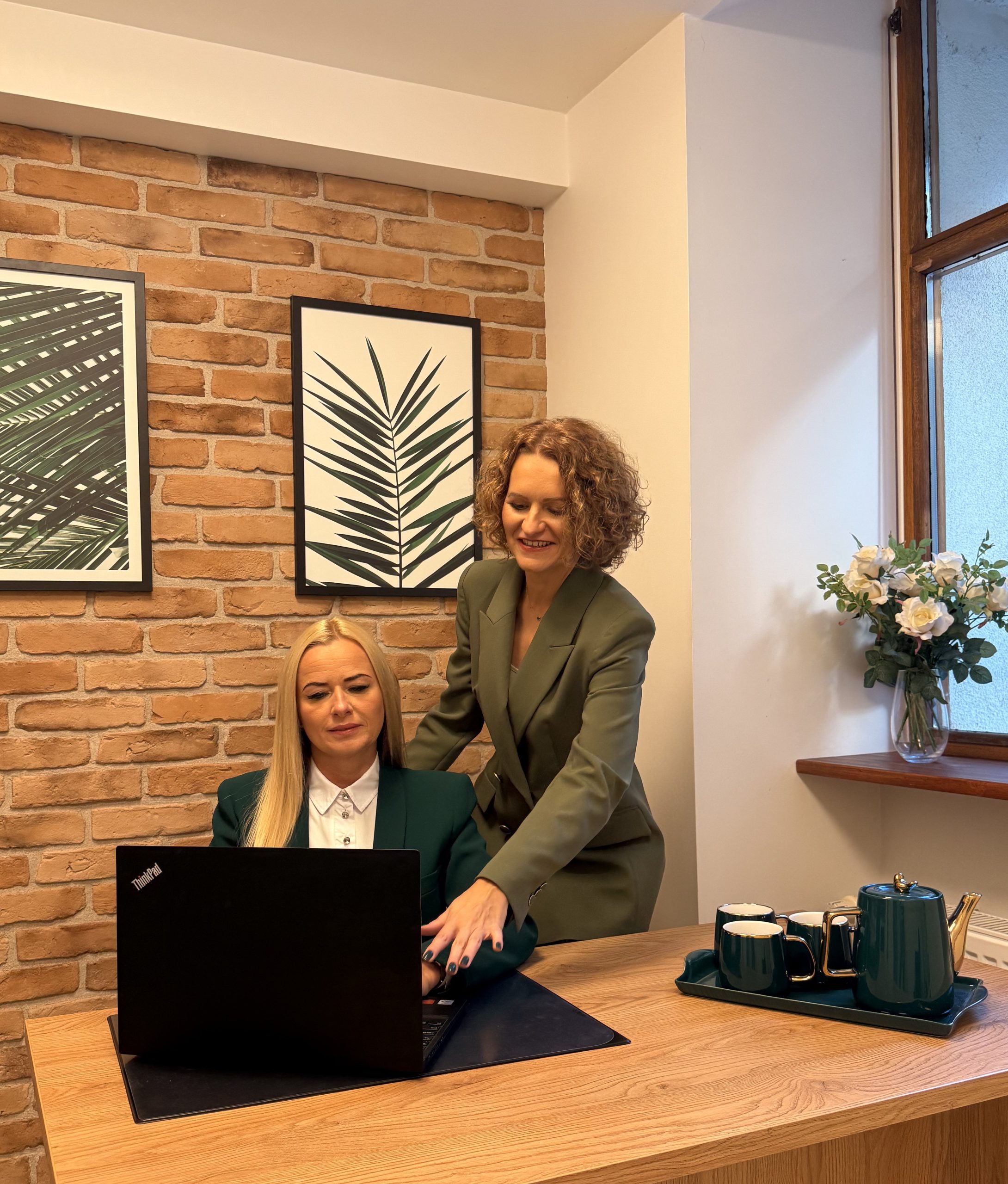 Dwie kobiety w zielonych marynarkach biznesowych w biurze. Jedna, siedząca z blond włosami, pracuje na laptopie, a druga, stojąca z kręconymi włosami, pochyla się nad nią i wskazuje na ekran. W tle ceglana ściana i obrazy z motywem liści.